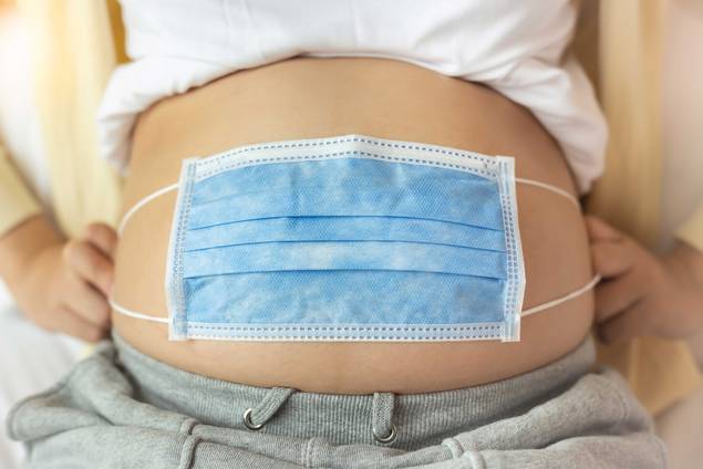 Eine Nahaufnahme eines schwangeren Bauches mit einer blauen chirurgischen Maske darüber, die Gesundheit und Sicherheit während der Schwangerschaft symbolisiert. Die Person trägt eine graue Hose und hat die Hände auf die Seite gestützt.