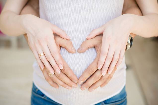 Eine schwangere Frau und ihr Partner stehen dicht beieinander und bilden mit ihren Händen über ihrem Bauch eine Herzform. Beide tragen Eheringe, die Frau ist mit einem weißen Oberteil und Jeans bekleidet.