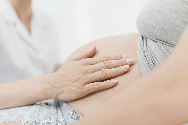 Eine Nahaufnahme des Bauches einer schwangeren Person, auf den eine medizinische Fachkraft während einer medizinischen Untersuchung sanft ihre Hand legt.