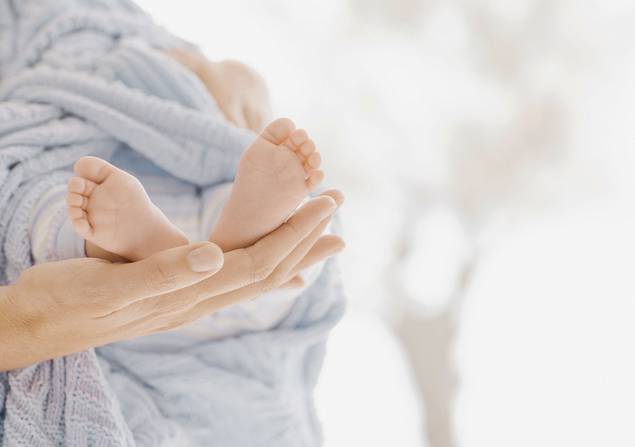 Nahaufnahme einer erwachsenen Hand, die sanft die nackten Füße eines in eine weiche, hellblaue Decke eingewickelten Babys streichelt, mit einem sanft verschwommenen Hintergrund.