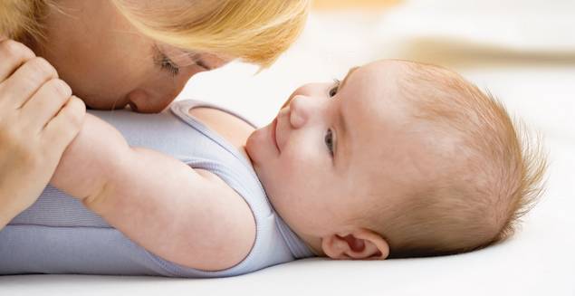 Eine Frau mit blondem Haar küsst sanft die Nase eines lächelnden Babys, das auf dem Rücken liegt, beide auf einer weichen weißen Unterlage. Das Baby schaut die Frau an, die einen hellblauen ärmellosen Strampler trägt.