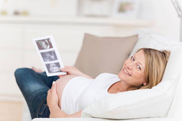 Eine schwangere Frau liegt auf einer weißen Couch, lächelt in die Kamera, hält Ultraschallbilder in einer Hand und berührt mit der anderen ihren Bauch. Sie trägt ein weißes Hemd und eine Jeans.